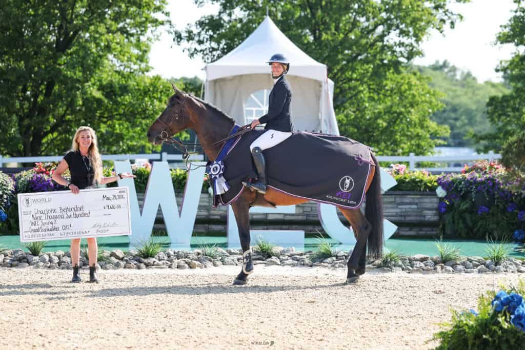A rider on a horse dressed in competition attire poses with a rosette, while a woman holds a large winner's check outdoors near a white tent and decorative plants.