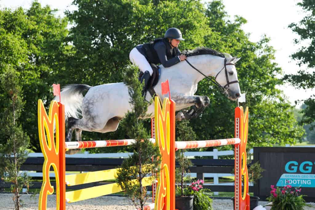 A rider wearing a helmet and formal attire jumps a gray horse over a brightly colored obstacle in an outdoor equestrian event.
