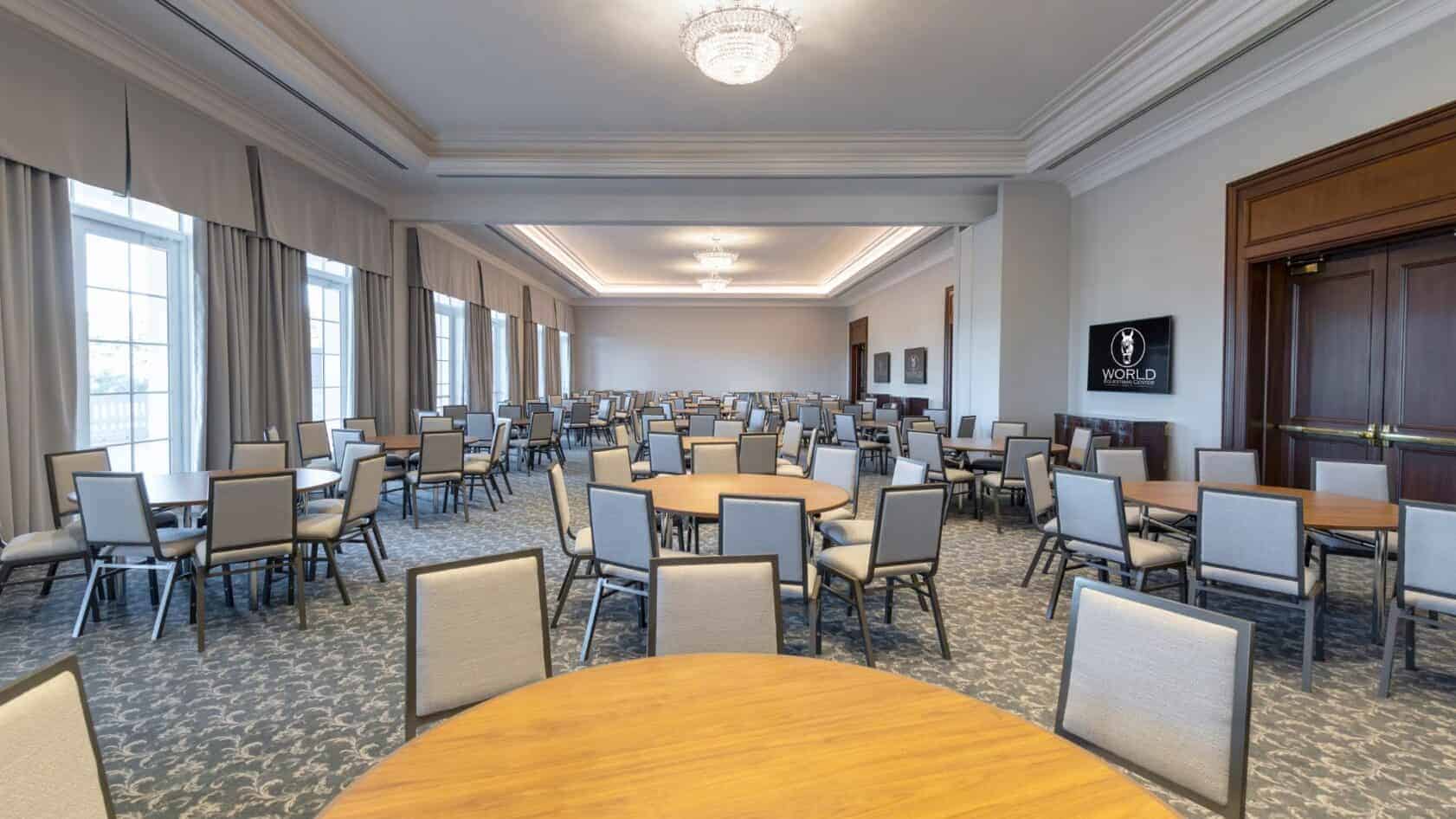 A spacious conference room with round tables and many chairs arranged neatly, large windows on one side, and a screen displaying the word "WORLD" on the wall.