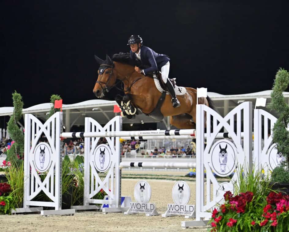An equestrian athlete rides a horse over a show jumping obstacle at an outdoor arena during a nighttime competition.