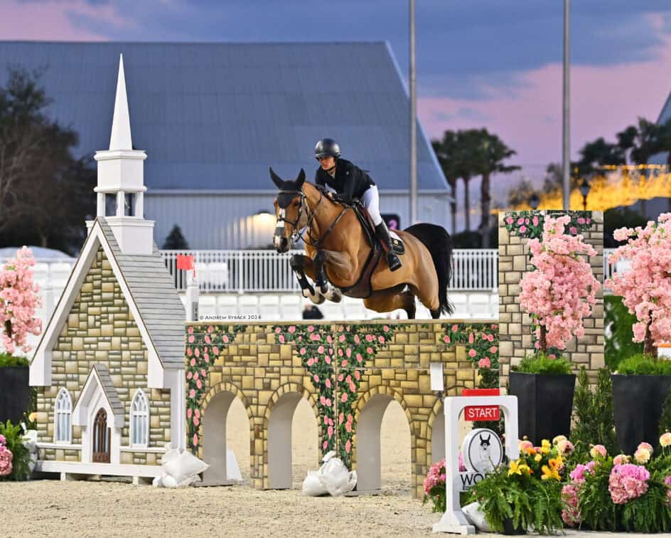 A rider on a horse jumps over an obstacle decorated like a stone arch and chapel, surrounded by pink flowers, at an equestrian event.