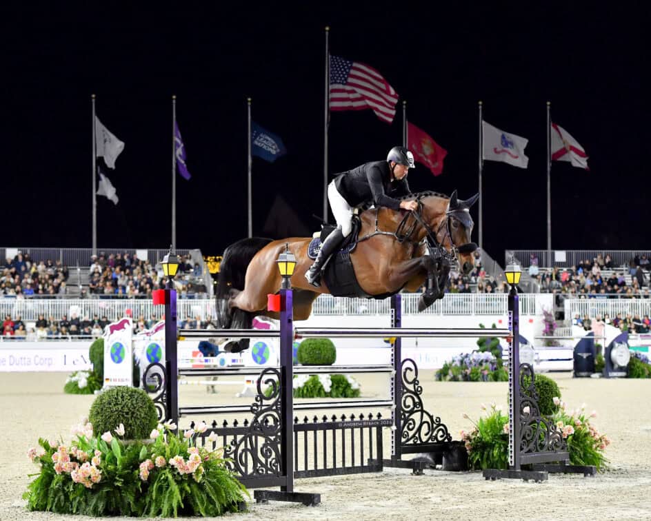 Equestrian rider and horse jumping over a high obstacle during a nighttime competition, with spectators and flags in the background.