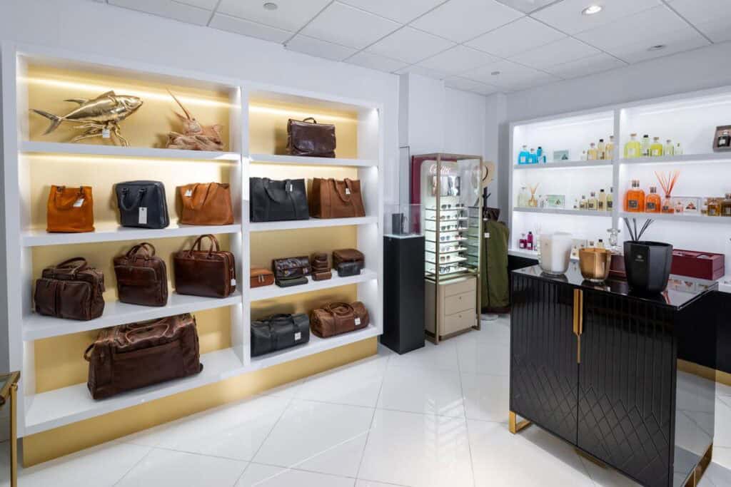 A retail display features shelves with leather bags, wallets, and accessories on the left, and shelves with perfumes and cosmetics on the right in a brightly lit store.