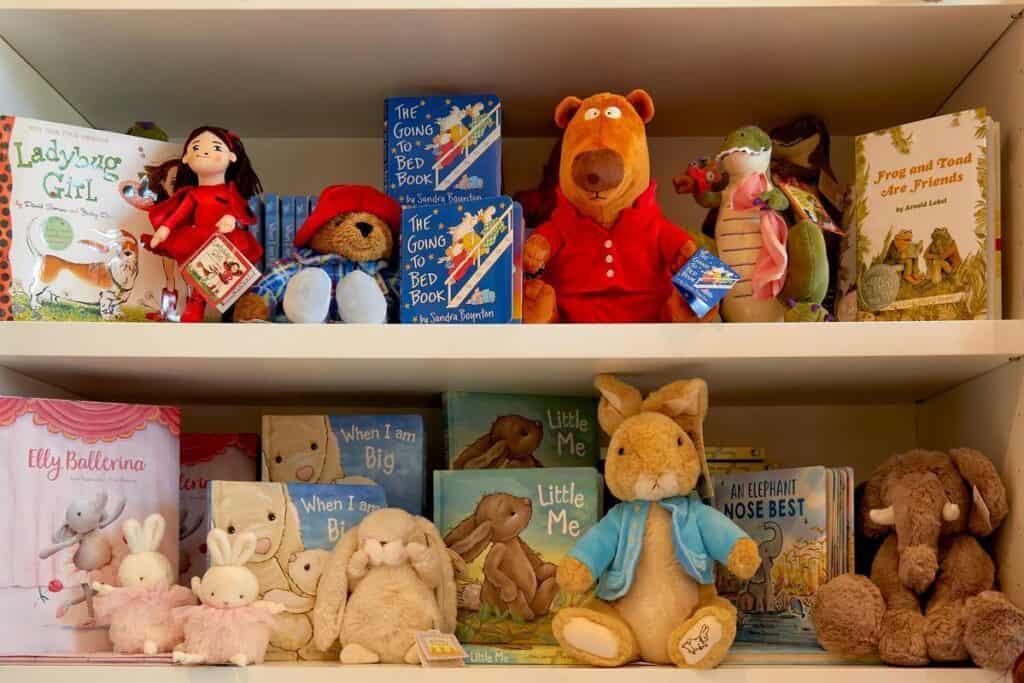 Three shelves display a variety of children's stuffed animals and dolls, arranged alongside colorful children's books with illustrated covers.