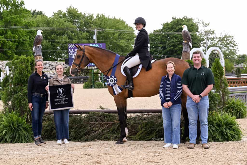 A rider on a horse wearing a ribbon poses with four people at an outdoor equestrian event; one person holds a sign with event details.