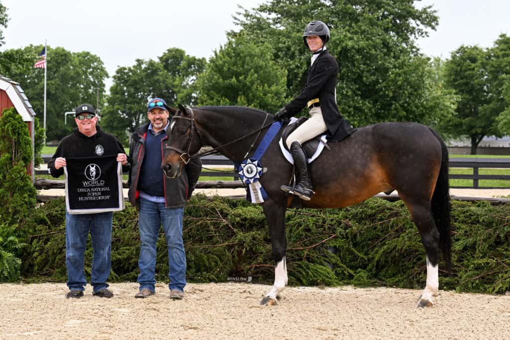 A rider on a horse with a winner's ribbon poses with two men holding a sign at an outdoor equestrian event. Trees and a fence are in the background.