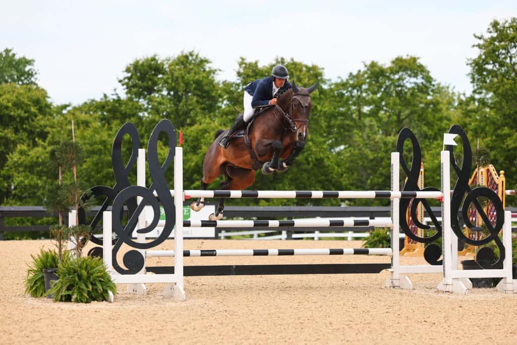 A rider on horseback jumps over a black-and-white obstacle decorated with treble clef symbols during an outdoor equestrian event.