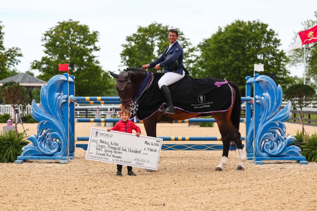 A man in riding attire sits on a horse, while a child stands holding a large winner's check. They are in an outdoor arena with blue decorative jumps in the background.