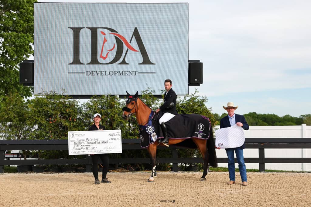 A rider on horseback holds a ribbon, flanked by two people, one holding a large check and the other a saddle pad, in front of an IDA Development sign at an outdoor event.