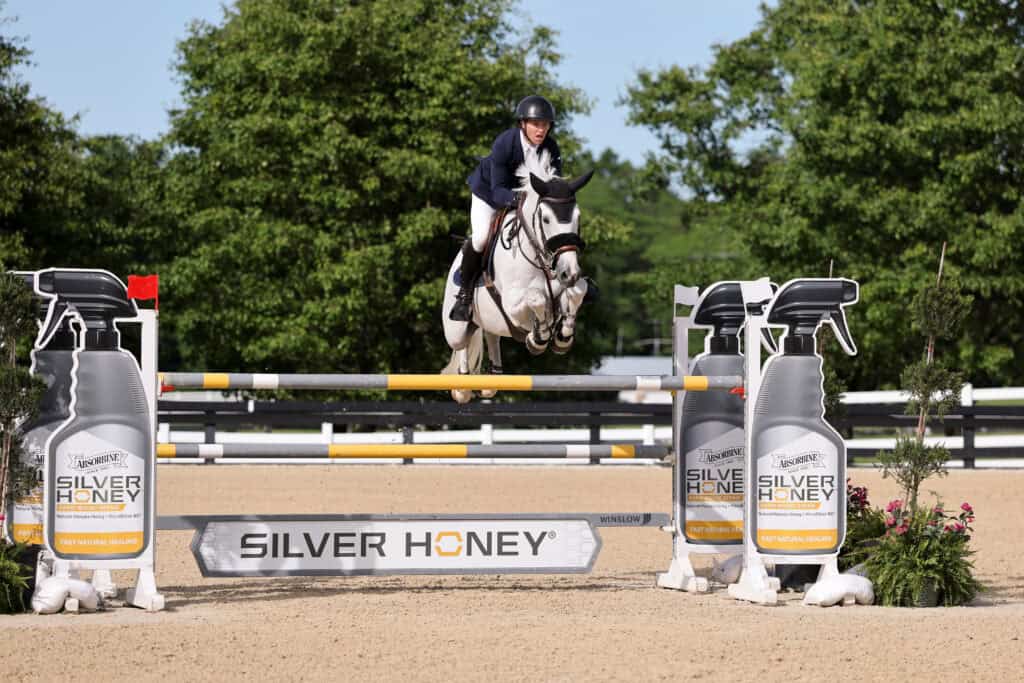 A rider on a white horse jumps over an obstacle labeled "SILVER HONEY" during an equestrian event on a sunny day.