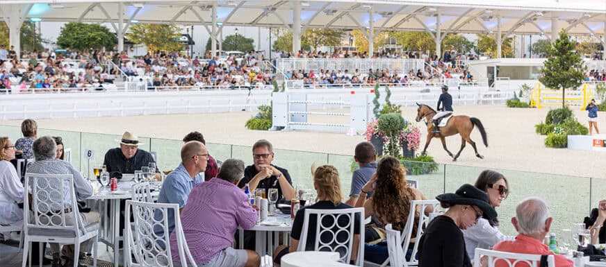 People dine at outdoor tables overlooking an equestrian arena, where a rider on horseback is performing in front of a seated crowd under a covered structure.
