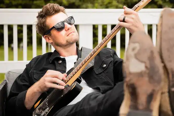 Man wearing sunglasses and a black jacket reclines on a couch with his feet up, playing an electric guitar outdoors.