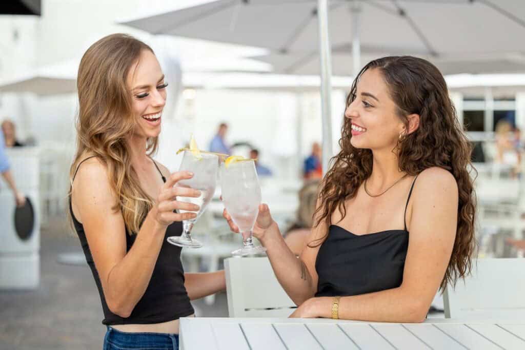 Two women sitting at an outdoor table, smiling and clinking glasses of water garnished with lemon slices.