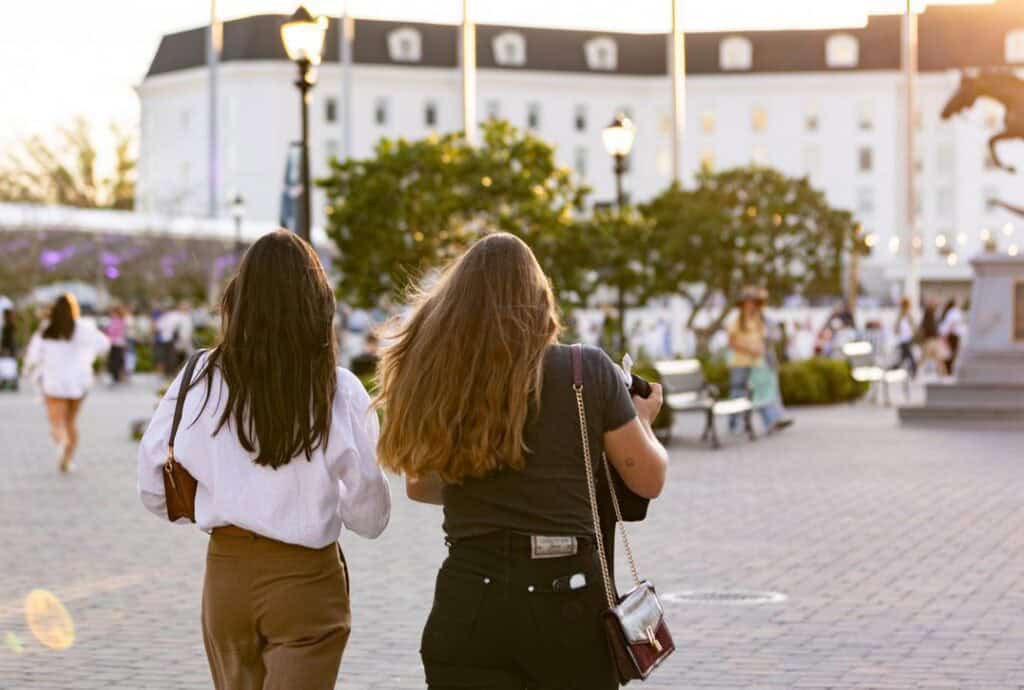Two women walk side by side on a paved plaza with trees, benches, and a white building in the background during sunset.