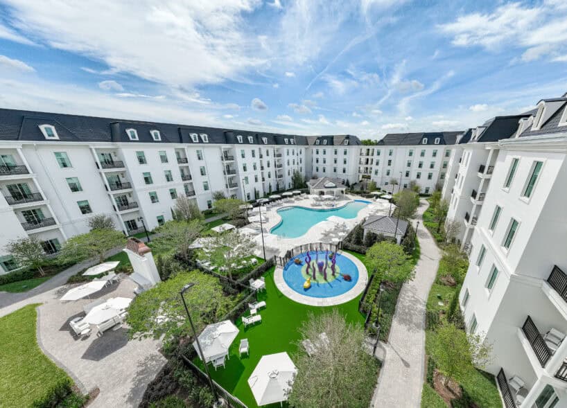 Aerial view of a modern apartment complex with a central outdoor pool, lounge areas, and a circular children's splash pad, surrounded by landscaped greenery and white buildings.