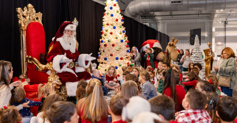 Santa Claus sits on a red throne, addressing a group of children gathered in front of him, with decorated Christmas trees and festive decorations in the background.