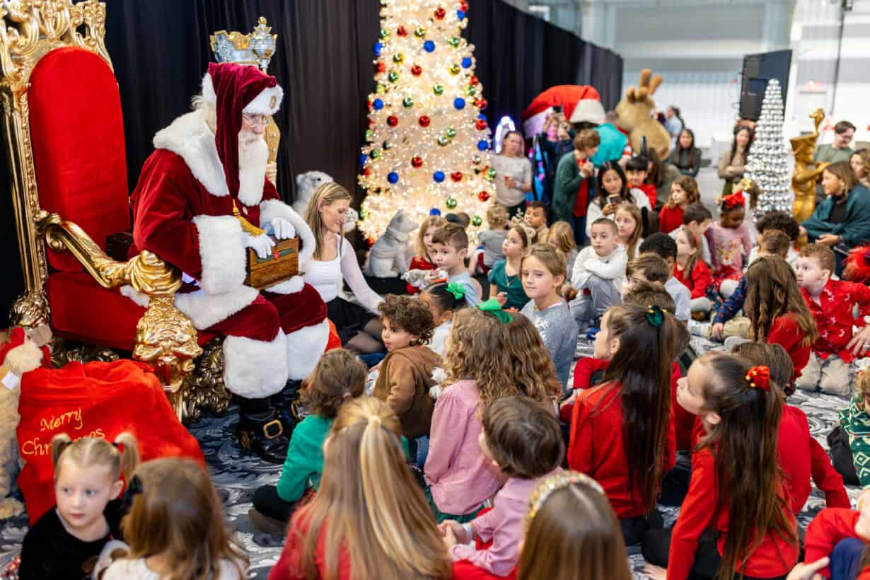 A person dressed as Santa Claus sits on a throne, speaking to a large group of children gathered in front, with festive decorations and a Christmas tree in the background.