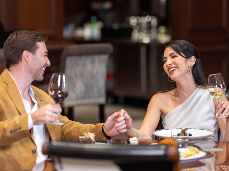 A man and woman sit at a dining table, holding hands and smiling while toasting with wine glasses in a restaurant setting.