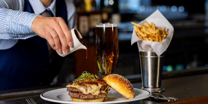 A server pours sauce over a cheeseburger on a plate beside a glass of beer and a metal cup of French fries at a bar counter.