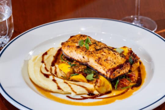 A plate of grilled salmon on a bed of vegetables and mashed potatoes, garnished with microgreens and drizzled with sauce.