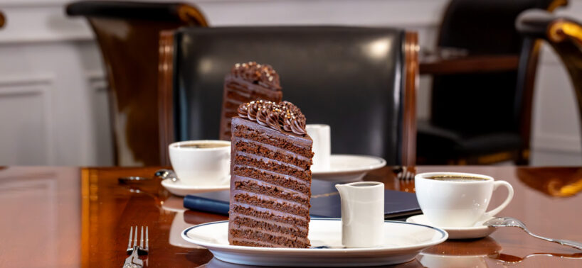 Two slices of layered chocolate cake on plates with cups of coffee and utensils, set on a polished wooden table in a formal dining setting.