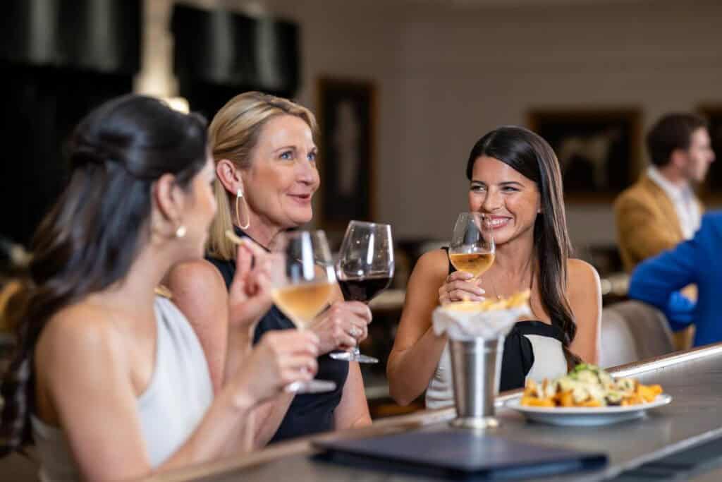 Three women sit at a bar holding wine glasses and talking, with a plate of food in front of them and other people in the background.
