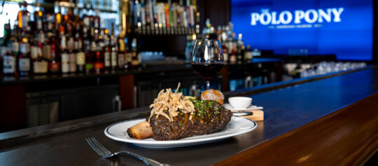 A steak topped with fried onions, a bread roll, sauce, and a glass of red wine are served on a bar counter with a "Polo Pony" sign in the background.