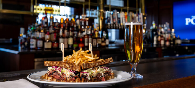 A plate with a stacked sandwich and fries sits on a bar counter next to a glass of beer, with bottles and taps visible in the background.