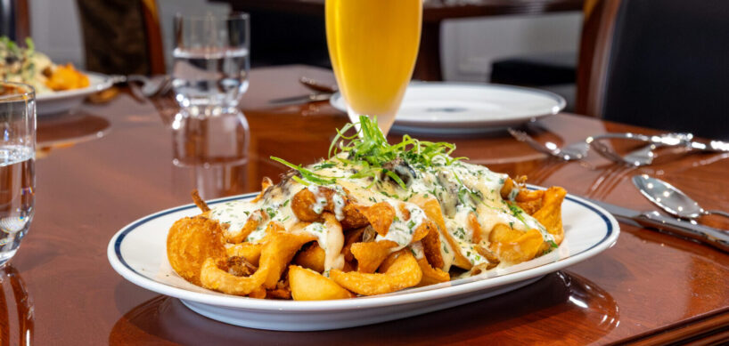 A plate of loaded potato wedges and a tall glass of beer on a wooden table in a restaurant, with a dog painting on the wall in the background.