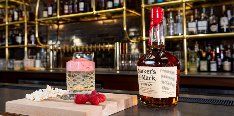 A bottle of Maker’s Mark whiskey next to a cocktail with foam, raspberries, and popcorn on a wooden board, set on a bar counter with liquor bottles in the background.