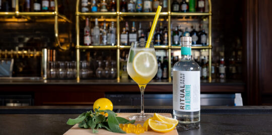 A glass of gin cocktail with lemon slices, yellow straw, and garnish sits on a bar counter beside a bottle labeled "Ritual Gin Alternative" and a cutting board with lemons and herbs.