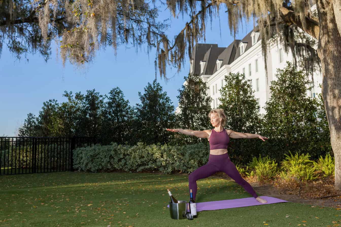 A woman in a purple workout outfit practices yoga in warrior pose on a mat outside, with trees and a large building in the background.