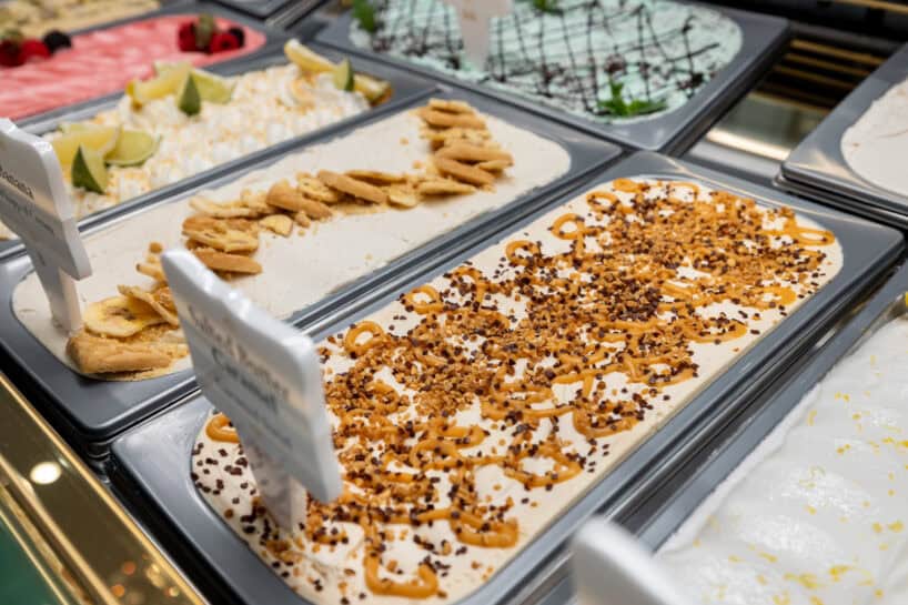 Several trays of assorted gelato flavors are displayed in a glass case, each decorated with various toppings such as pretzels, banana slices, lime, and chocolate drizzle.