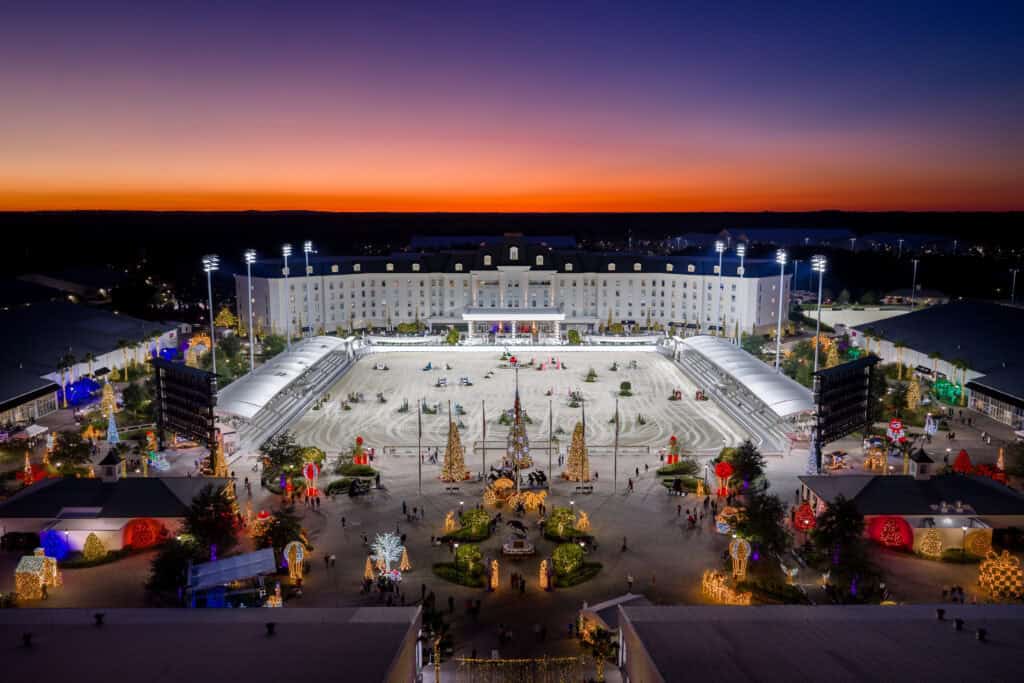 A large illuminated arena with decorated trees and holiday lights is shown at dusk, with a grand building in the background and the sky glowing orange and purple.
