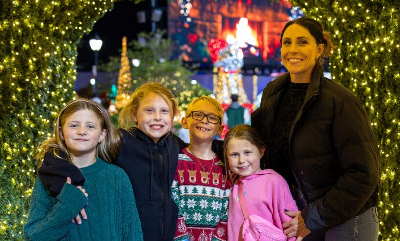 A woman and four children stand on a red carpet under a tunnel of holiday lights, posing for a photo and smiling at the camera.