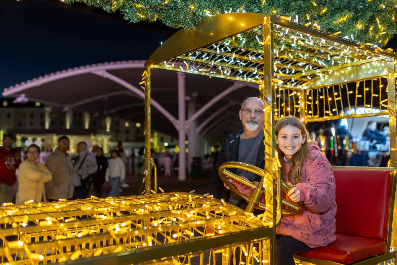 A man and a young girl sit on a decorative vehicle covered in holiday lights at an outdoor event, with people and festive lighting in the background.
