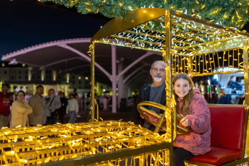 A man and a young girl sit on a decorative vehicle covered in holiday lights at an outdoor event, with people and festive lighting in the background.