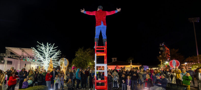 A performer in a red jacket stands atop a tall stack of chairs with arms outstretched, facing a nighttime crowd gathered in an outdoor plaza decorated with holiday lights.