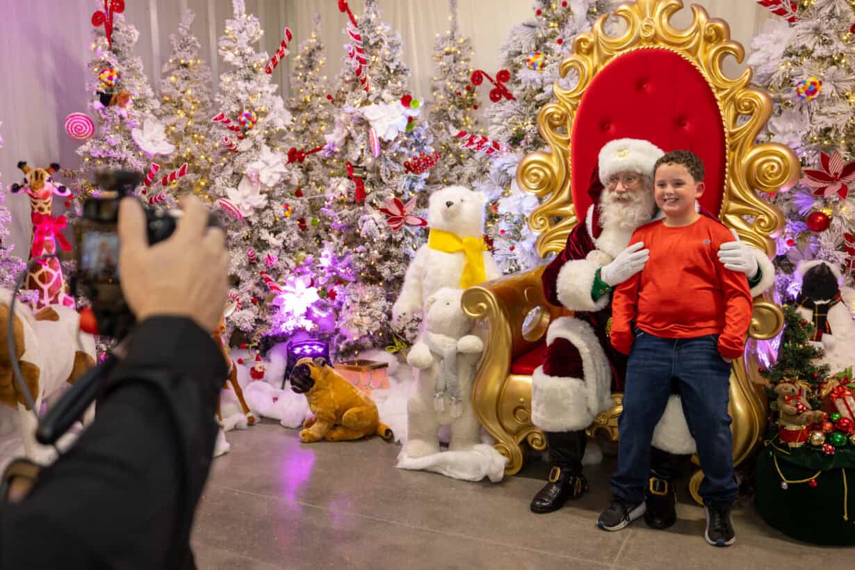 A boy in a red shirt poses with Santa Claus on a golden throne, surrounded by decorated Christmas trees and stuffed animals, as someone takes their photo.