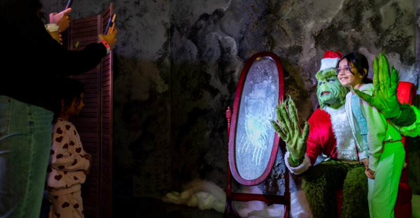 A child poses for a photo with a person dressed as the Grinch in front of a mirror, while two people take pictures.