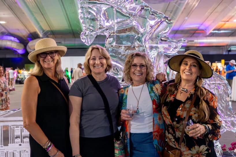 Four women standing together and smiling in front of an ice sculpture at an indoor event; three are holding drinks and dressed in casual clothing and hats.