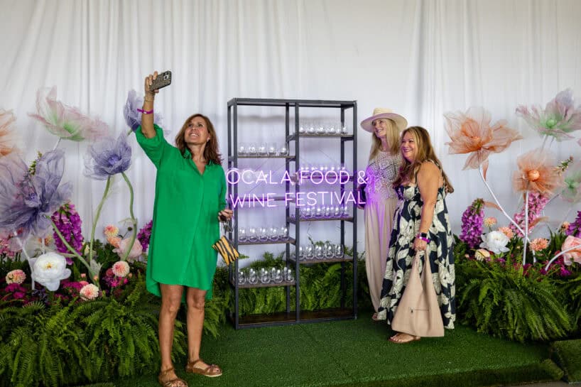 Three women pose for a selfie in front of a neon "Ocala Food & Wine Festival" sign, surrounded by large artificial flowers and greenery.