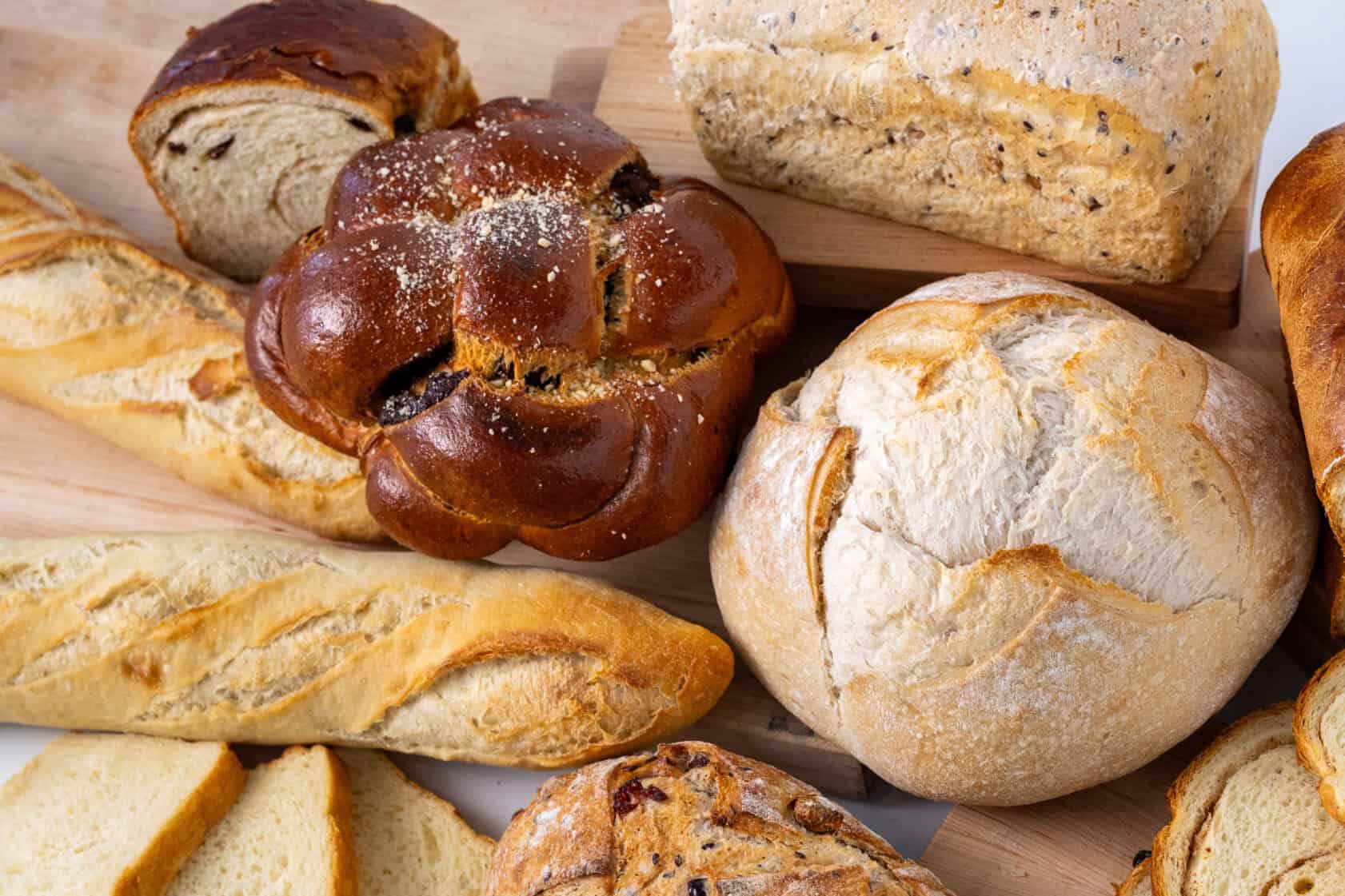 An assortment of various breads, including baguettes, a round loaf, braided bread, sliced bread, and a multigrain loaf, arranged on wooden boards.