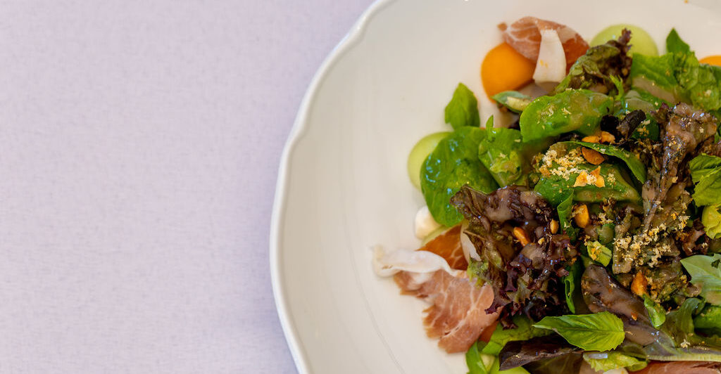 A white plate with a mixed green salad, slices of prosciutto, melon balls, and sprinkled nuts on a light tablecloth.