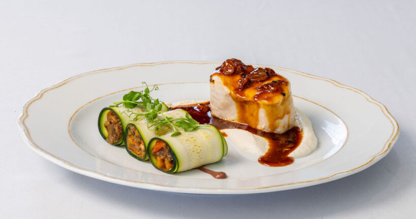 A plated dish with a piece of fish topped with brown sauce, served alongside three zucchini rolls filled with vegetables and a white sauce, garnished with microgreens.