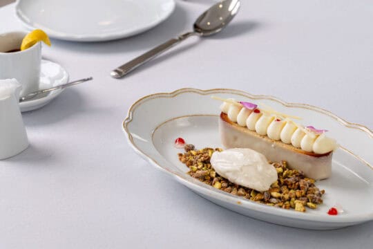 A plated dessert with a rectangular pastry topped with cream, served with a scoop of ice cream and nut crumble on a white plate; utensils and a cup of tea in the background.