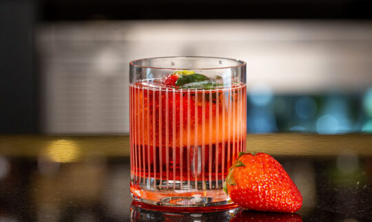 A glass of red strawberry cocktail with ice and mint sits on a reflective black surface, with a fresh strawberry placed beside the glass. The background is a blurred bar.