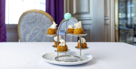 Six small appetizers topped with a white garnish are displayed on a tiered metal stand, set on a white tablecloth in an elegant dining room.