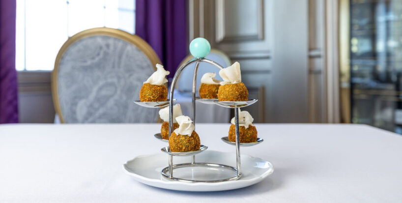 Six small appetizers topped with a white garnish are displayed on a tiered metal stand, set on a white tablecloth in an elegant dining room.