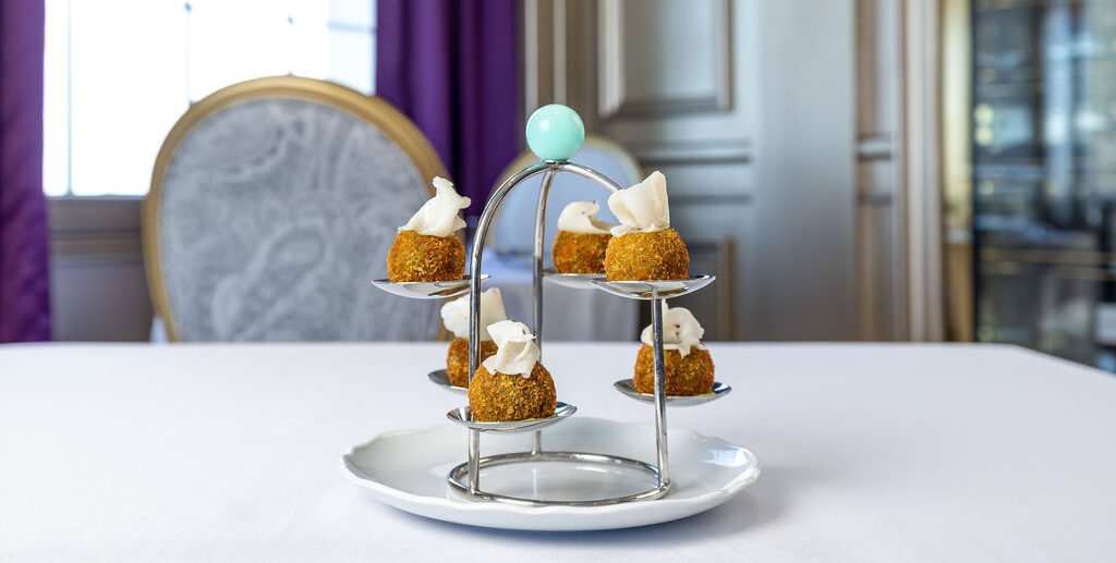 Six small appetizers topped with a white garnish are displayed on a tiered metal stand, set on a white tablecloth in an elegant dining room.